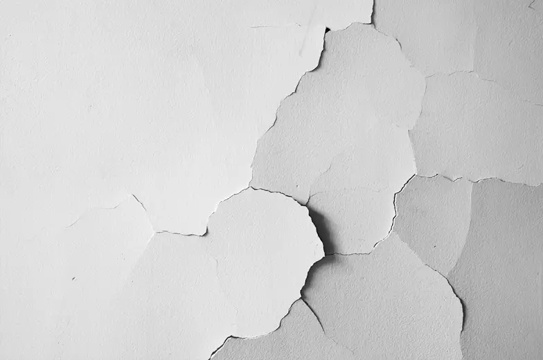 Das Bild zeigt eine Wand mit abgeblätterter weißer Farbe. Mehrere große und kleine Risse und Abplatzungen sind sichtbar, wobei die darunterliegende Oberfläche. Ein Schatteneffekt erzeugt Tiefenwirkung.