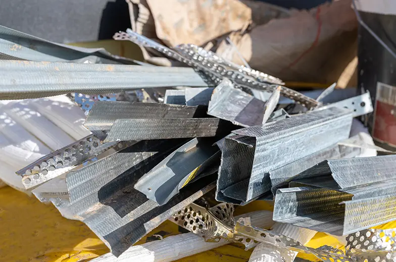 Ein Haufen von Metallprofilen und -streifen liegt auf einer gelben Oberfläche. Die Metallteile haben verschiedene Formen und Größen und sind teils perforiert, teils glatt. Im Hintergrund sind unscharfe Objekte erkennbar.